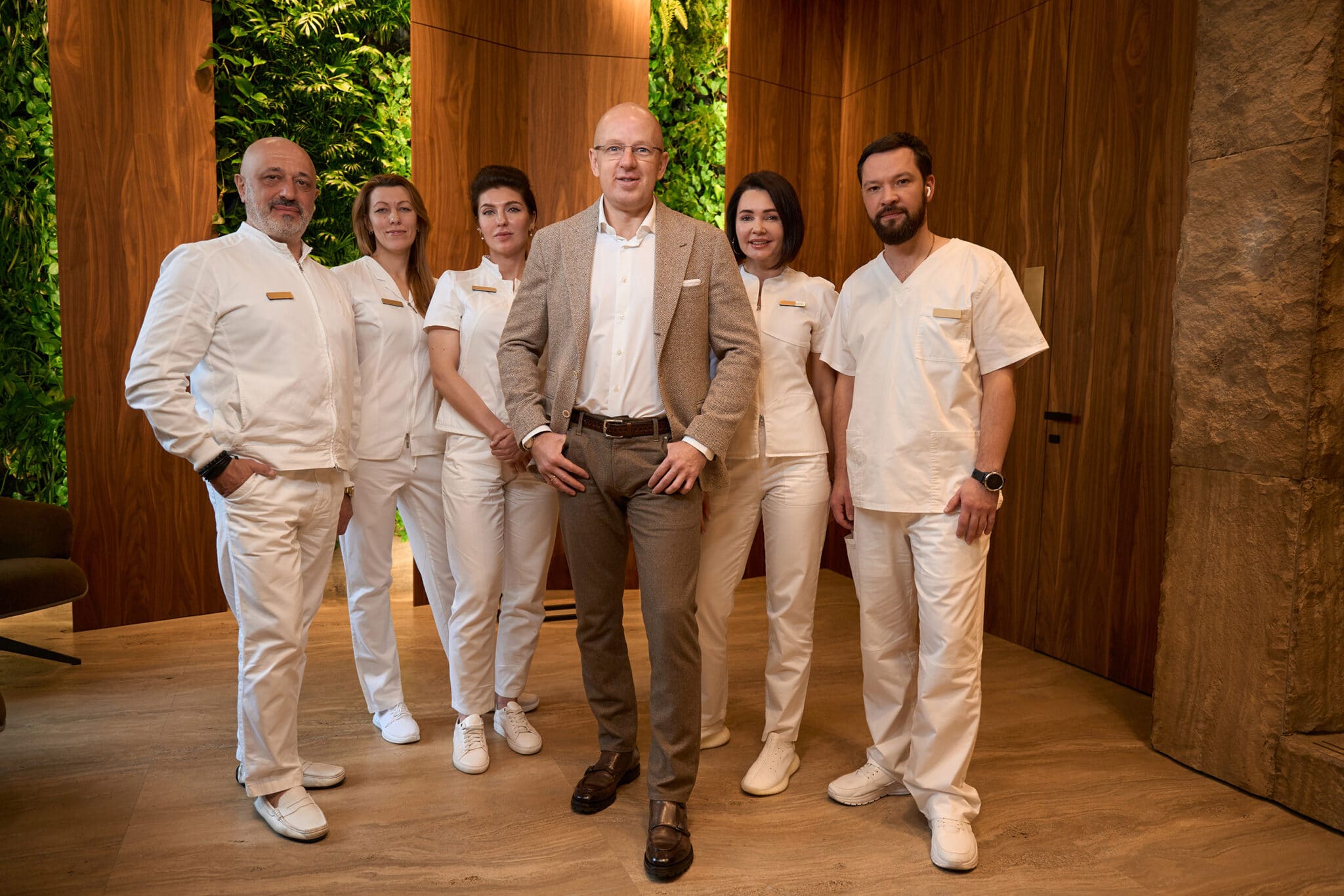 Group of people in medical clothes stands against background of a wooden wall, in center is man in business suit