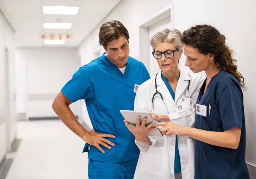 Mature female doctor discussing medical report with nurses in hospital hallway. Senior general practitioner discussing patient case status with group of medical staff after surgery. Doctor working on digital tablet while in conversation with healthcare workers, copy space.