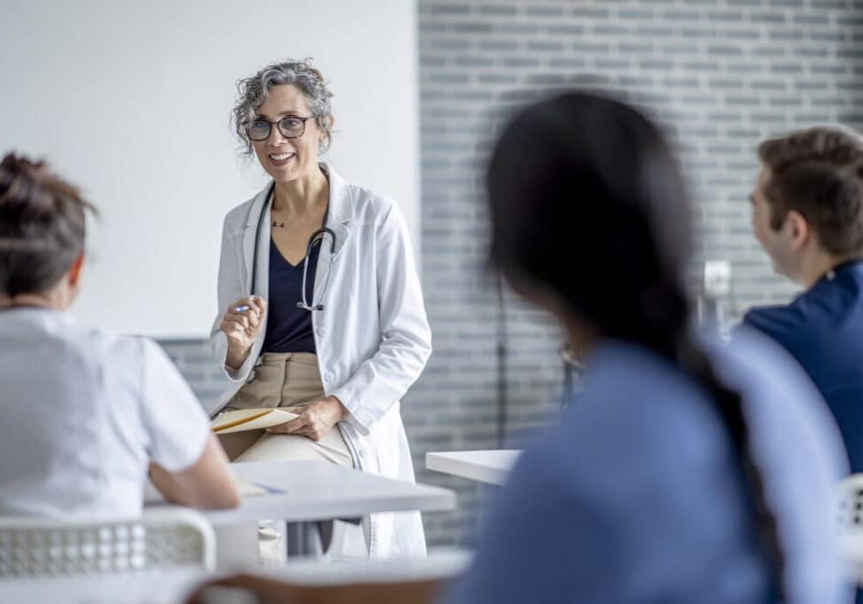 A mature female doctor sits at the front of a classroom as she teaches a small group of students.  She has a file folder on her lap as she talks with the students, and is dressed in a white lab coat.  The students are seated in front of her and paying close attention as they take notes.