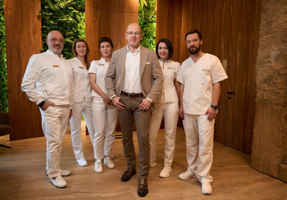 Group of people in medical clothes stands against background of a wooden wall, in center is man in business suit